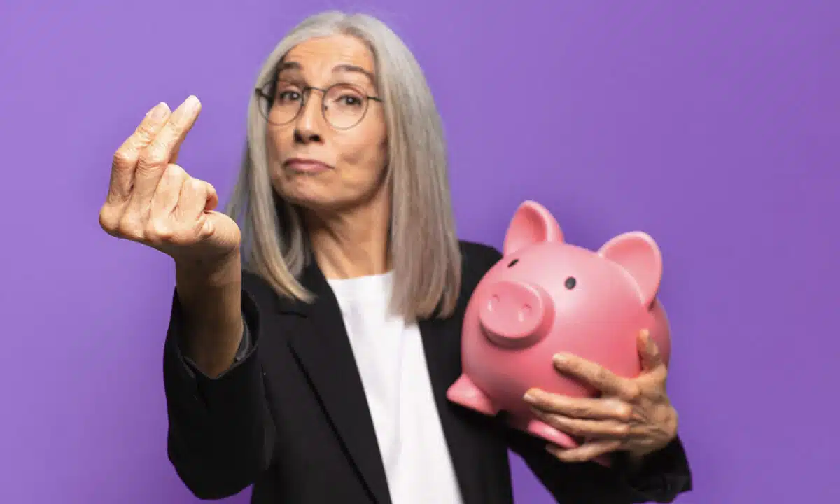 Femme senior qui tient une tirelire cochon et fait le signe d'argent avec ses doigts
