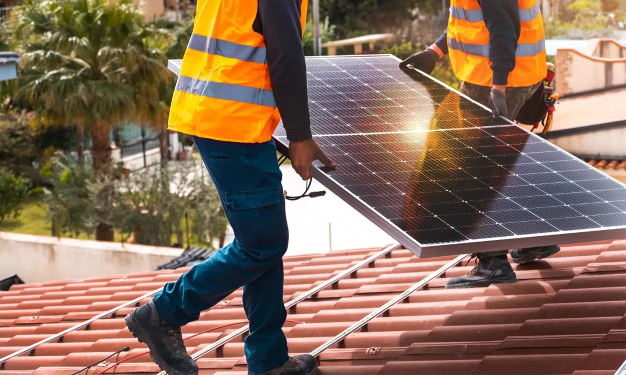 Deux installateurs de panneaux solaires portent un panneau photovoltaïque sur le toit d'une maison individuelle